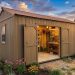 Garden shed with double doors open, revealing a well-organized workbench and tools inside, surrounded by blooming flowers in the yard at sunset.