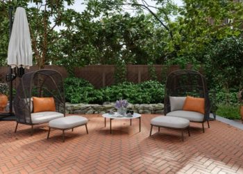 Two black wicker lounge chairs with light cushions and orange pillows on a brick patio, a small round table between them, and a closed umbrella nearby.