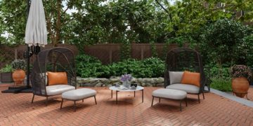 Two black wicker lounge chairs with light cushions and orange pillows on a brick patio, a small round table between them, and a closed umbrella nearby.