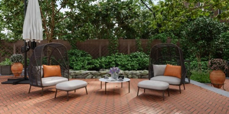 Two black wicker lounge chairs with light cushions and orange pillows on a brick patio, a small round table between them, and a closed umbrella nearby.