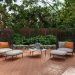 Two black wicker lounge chairs with light cushions and orange pillows on a brick patio, a small round table between them, and a closed umbrella nearby.