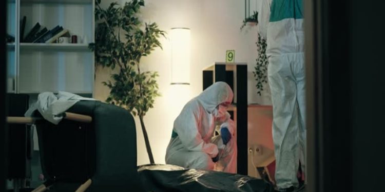 Two people in white hazmat suits examine a covered body bag on the floor in a clinical room with shelves and a lamp maybe visible in the background.